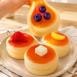 Hand stretching a sticky soft Blueberry Pancake Squishy Toy on a ceramic plate next to Strawberry, Orange, and Butter Souffle Pancake Squishies with removable toppings, perfect non-edible Christmas gift and stress relief sensory toy