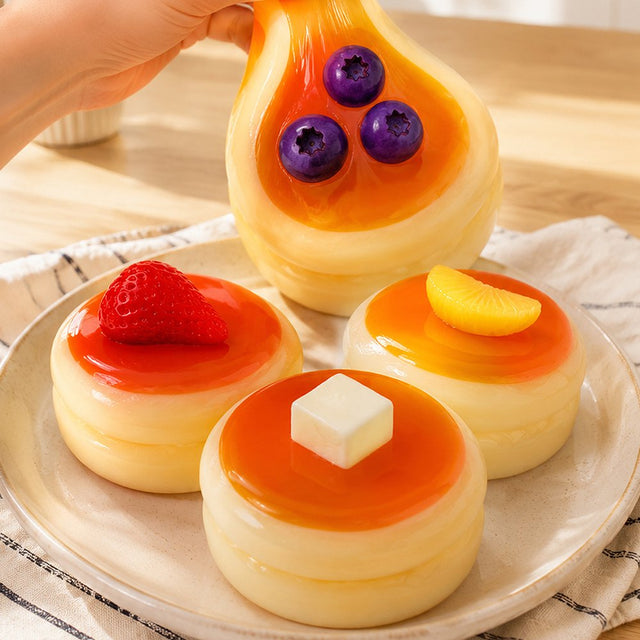 Hand stretching a sticky soft Blueberry Pancake Squishy Toy on a ceramic plate next to Strawberry, Orange, and Butter Souffle Pancake Squishies with removable toppings, perfect non-edible Christmas gift and stress relief sensory toy