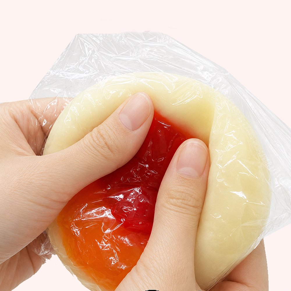 Hands pressing and kneading a soft sticky pancake squishy showing inner red jelly-like filling through clear protective film, demonstrating soft gel elasticity for stress relief and anxiety reduction