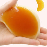 Hand holding and stretching the glossy syrupy top layer of a soft soufflé pancake squishy with an orange slice topping nearby, highlighting sticky TPR texture for sensory fidget lovers