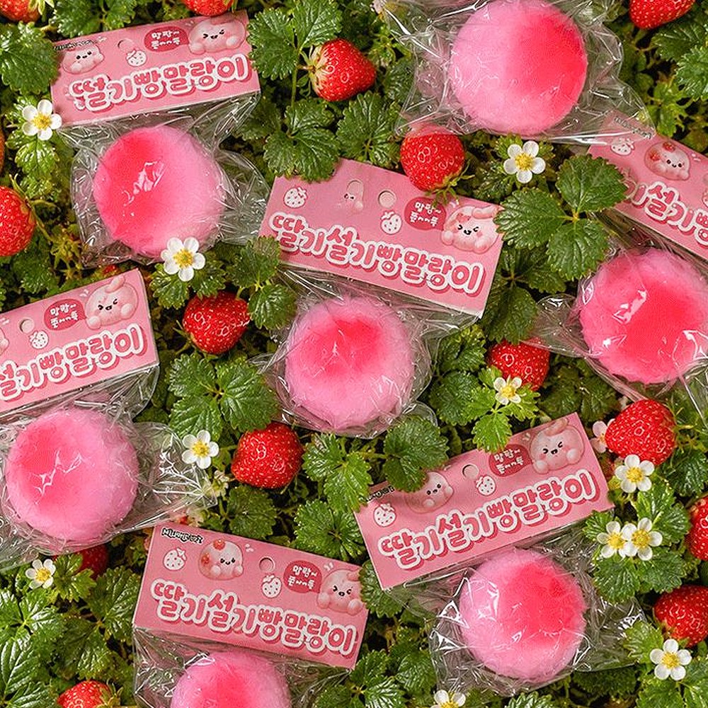 Multiple pink Strawberry Rice Cake Bread Squishies in Korean packaging scattered on green strawberry plants and white flowers, showcasing cute kawaii dessert fidget toys for anxiety relief and seasonal holiday gifting